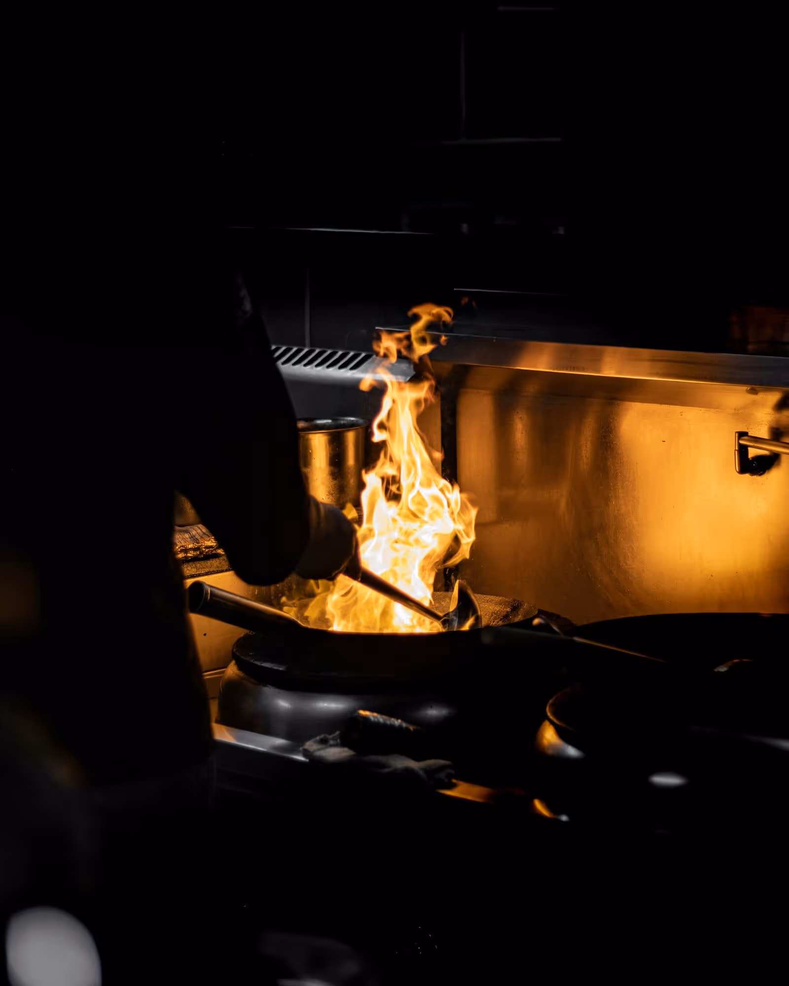 Flame jumping out of a pan on the range while a cook works the dark end of the line