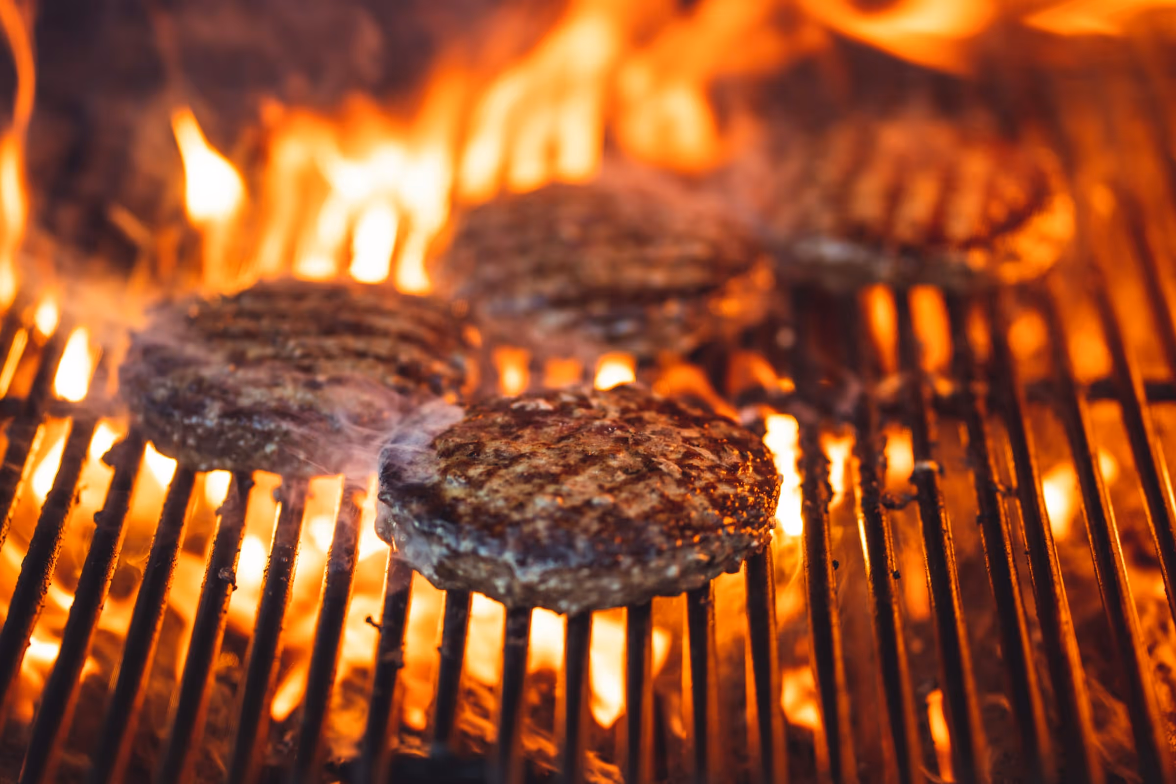 Beef patties searing on the grate over open flame with smoke coming off the crust