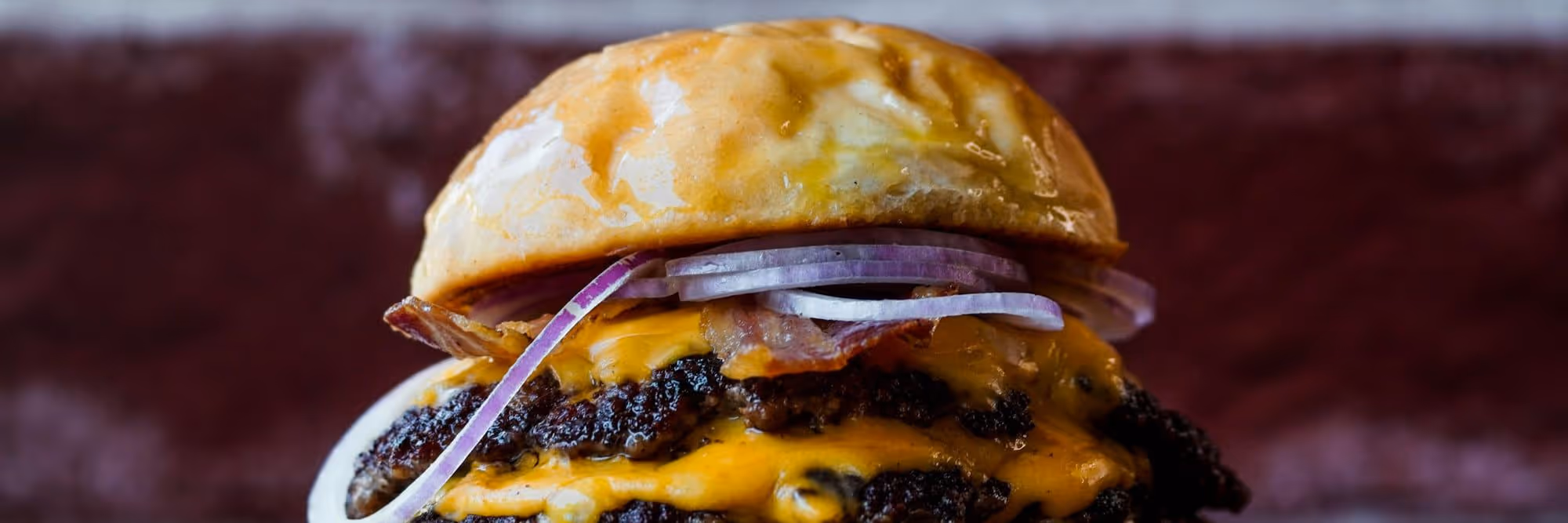 A double smash burger stacked with American cheese, bacon, and raw red onion under a hard rim light