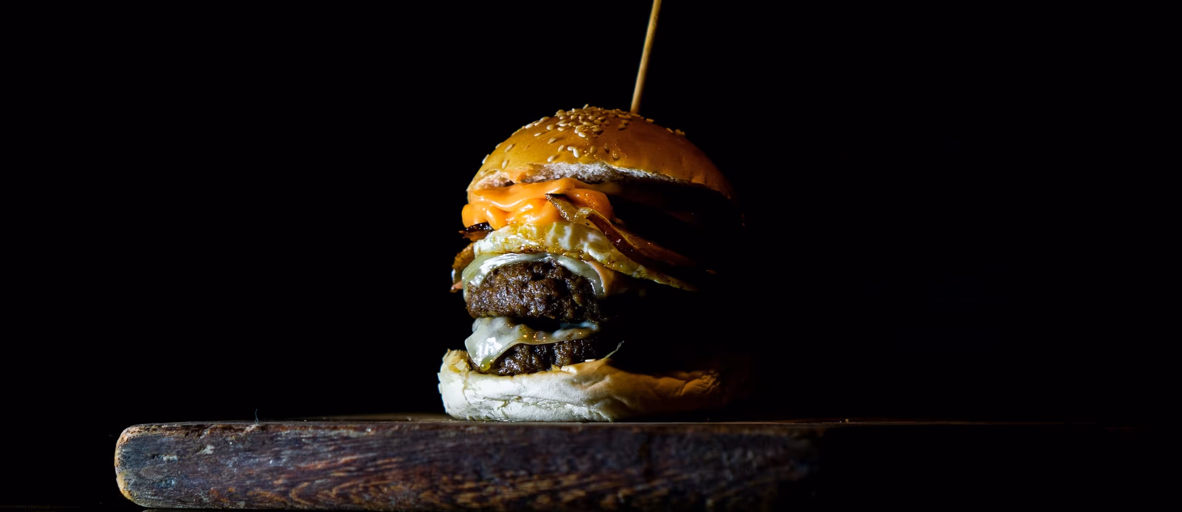A double smash burger with melted cheese and bacon standing on a worn wooden board in a black room