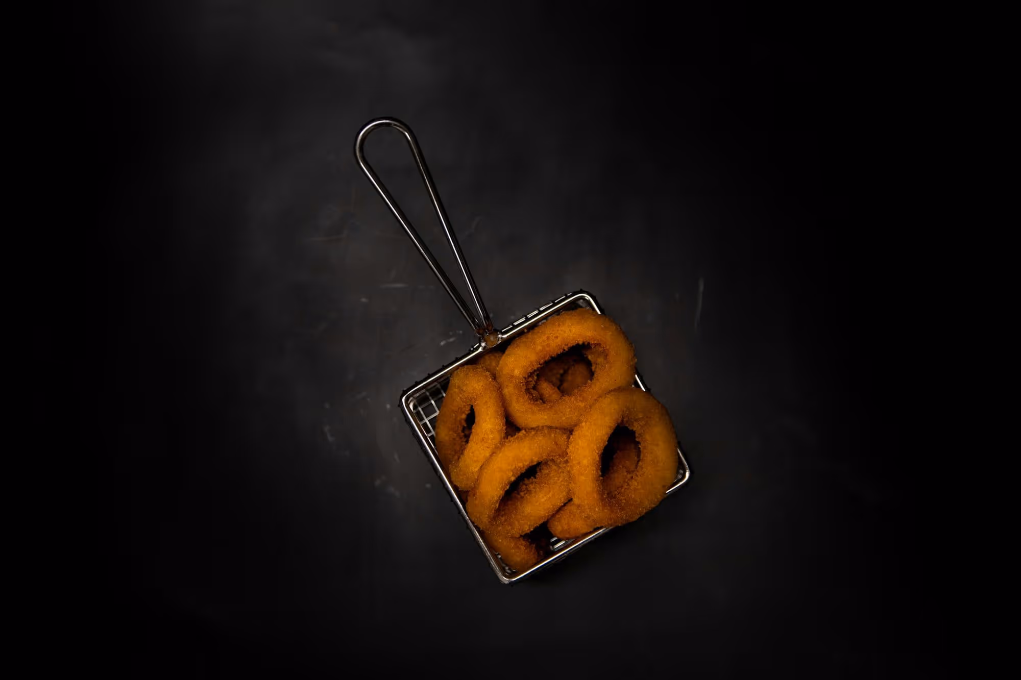 Six onion rings resting in a wire fry basket on the black steel of the pass