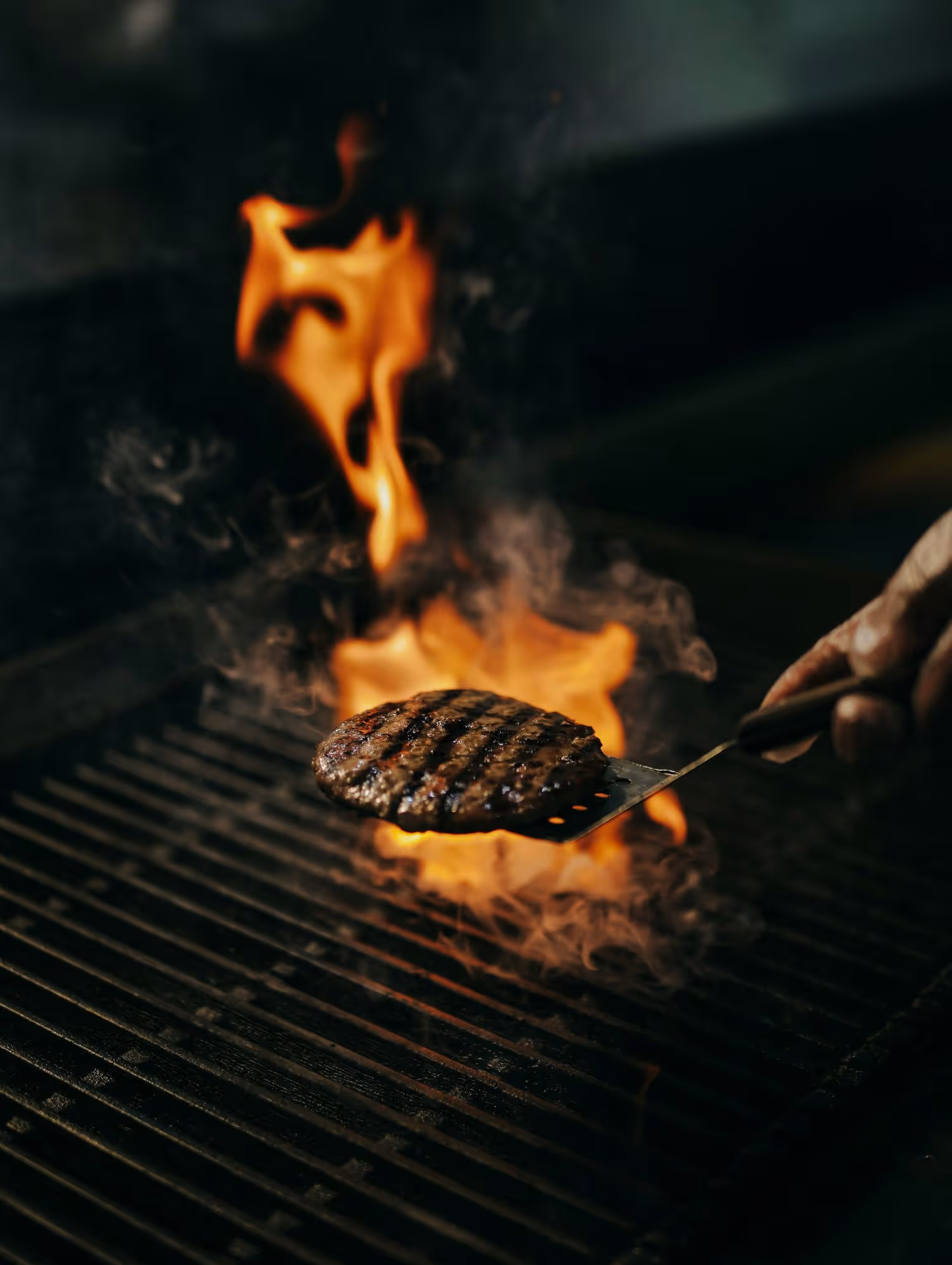 A patty lifted off the fire on a spatula with smoke coming off the grate